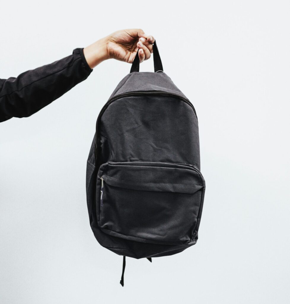 A simple black backpack being held by a hand against a minimal white background, emphasizing style and practicality.